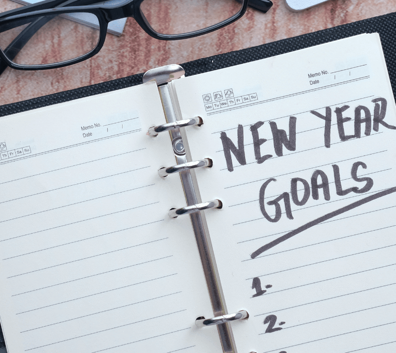 An open planner with NEW YEAR GOALS written at the top of a page, surrounded by glasses, a pen, and binder clips as part of planning 2026 trends and innovations.